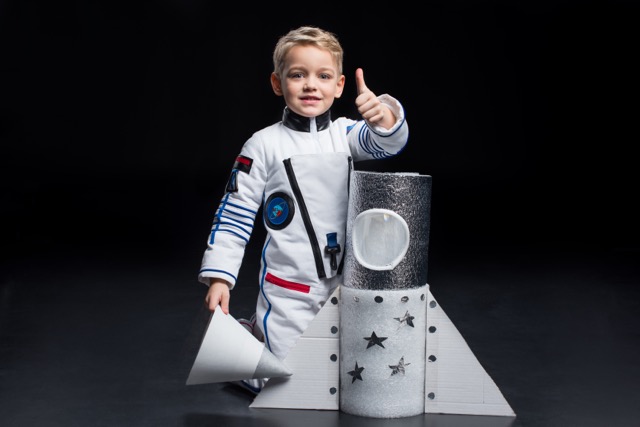 Smiling little boy astronaut in space suit kneeling near toy spaceship and showing thumb up